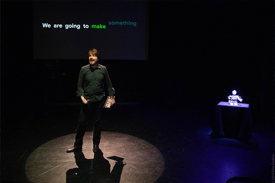 A performer of Artificial Intelligence Improvisation on stage with the words "we are going to make something" behind him.