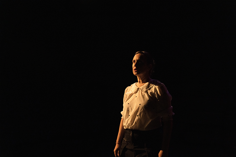 A woman stands on a dark stage lit dimly with a warm light, wearing a white blouse
