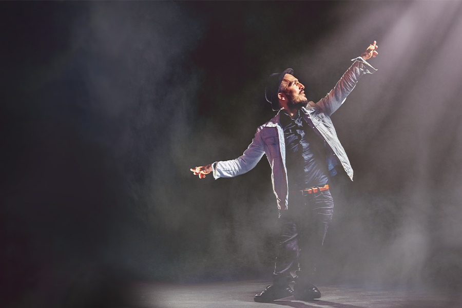A man stands on stage holding his hand in the air and gazing into it, the stage is filled with fog and lit with a spotlight