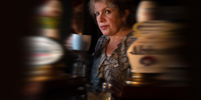 A blonde middle-aged woman drinking tea behind the bar in a traditional pub, across from her is a glass of whiskey