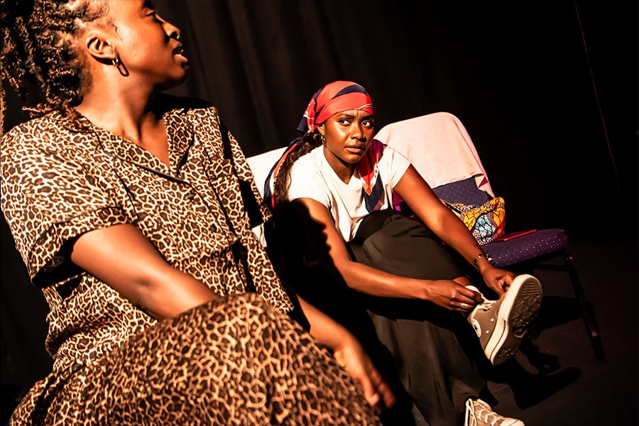 Two black women performing, one looks at the other disconcerted whilst tying the lace of her converse shoes up