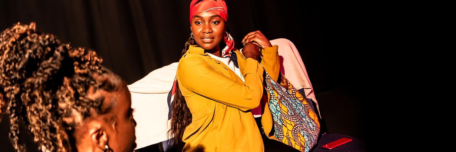 Two black women performing on stage. One is grabbing her bag and looking at the other.
