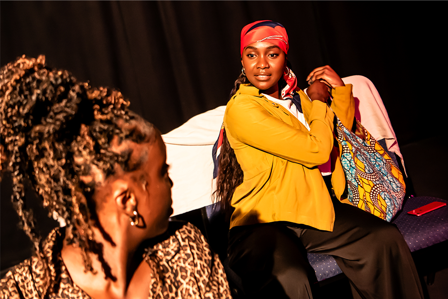 Two black women performing on stage. One is grabbing her bag and looking at the other.