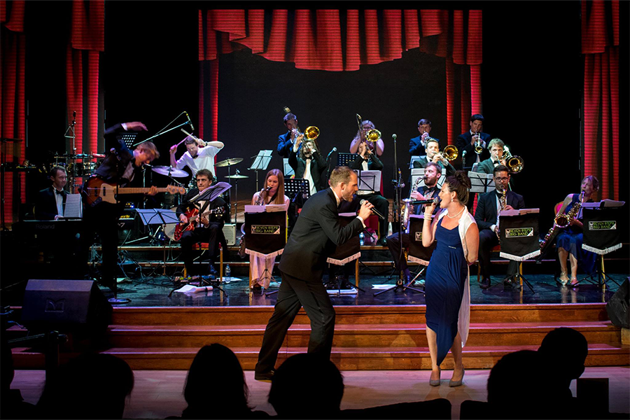 A male singer wearing a tux and a female singer wearing a navy dress sing to each other holding microphones accompanied by a band full of brass instruments