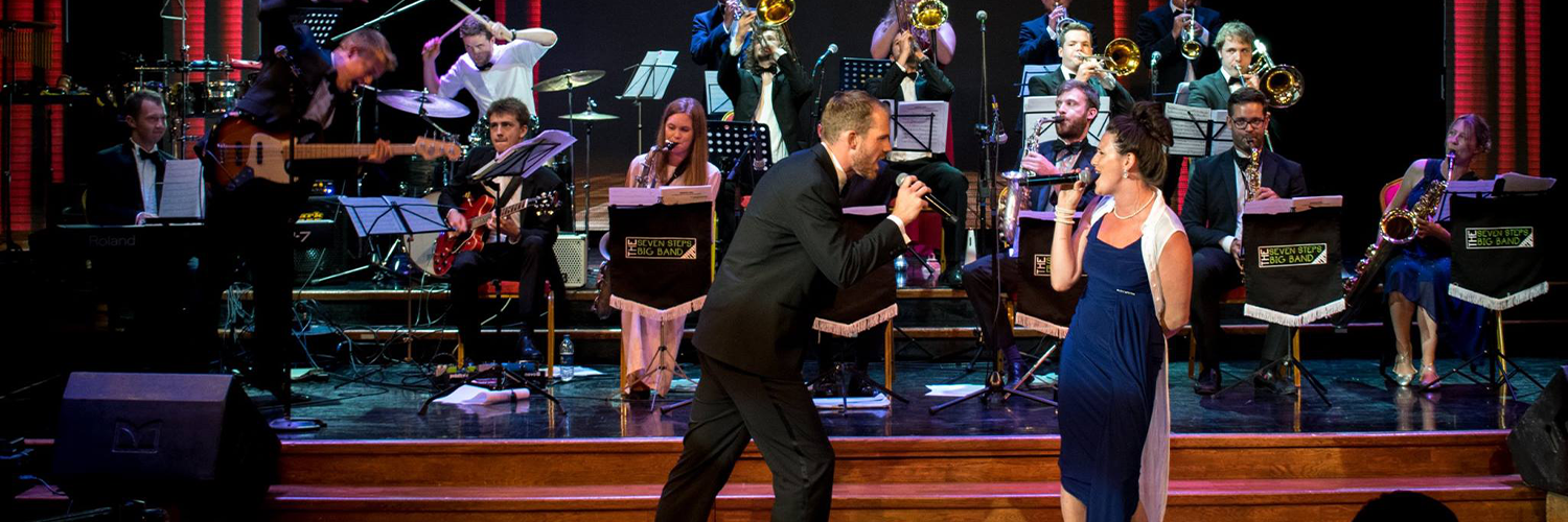 A male singer wearing a tux and a female singer wearing a navy dress sing to each other holding microphones accompanied by a band full of brass instruments