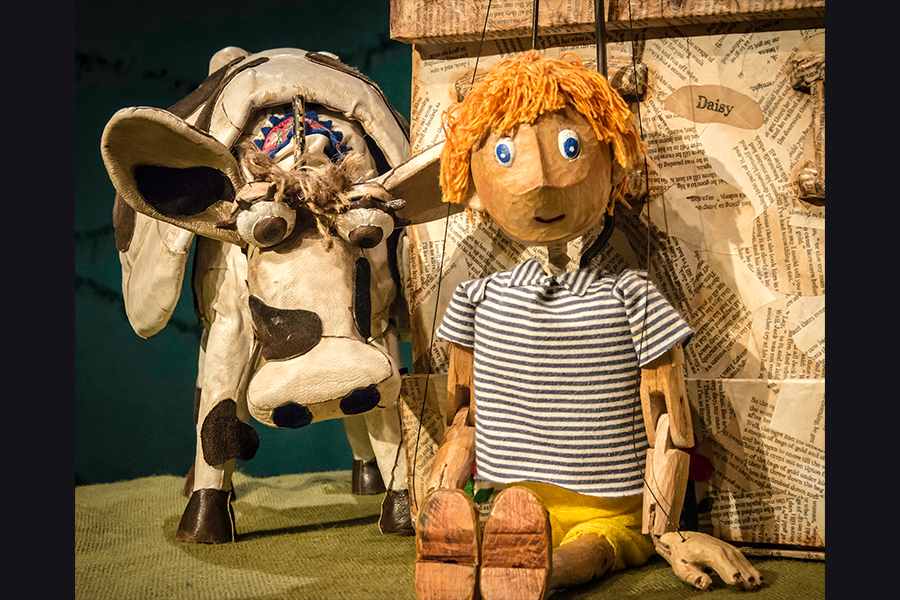 A boy puppet with ginger hair and blue eyes and a puppet cow sitting next to each other