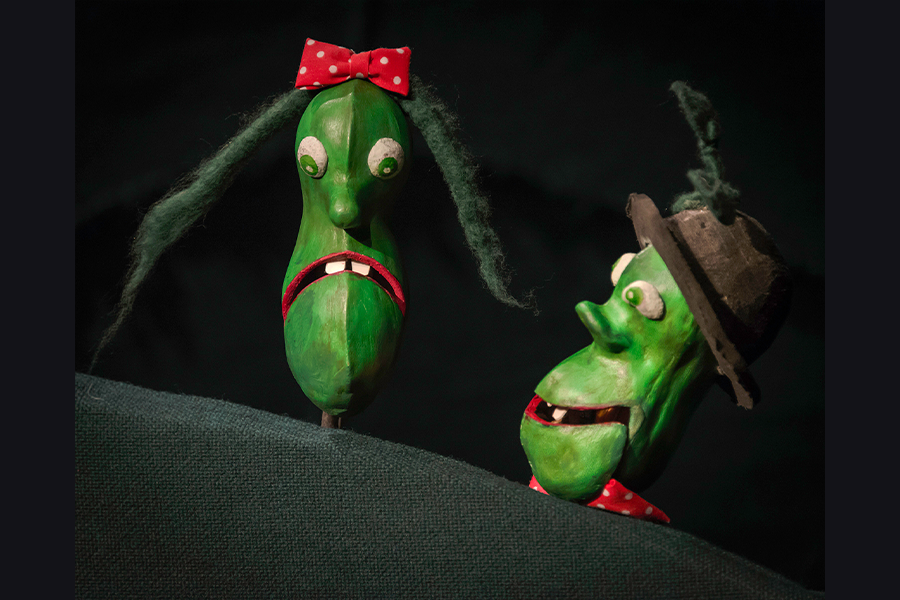 Two puppet green beans. One wears a red bow and the other wears a bowling hat.