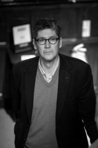 Black and white headshot of Martin. He is wearing a black blazer, a shirt and a jumper. He has short hair and wears glasses.