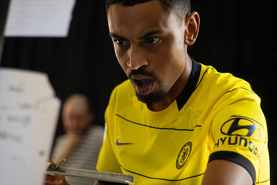 Tre Medley, a black actor wearing a neon yellow football top is staring intently at a piece of paper holding an ink block