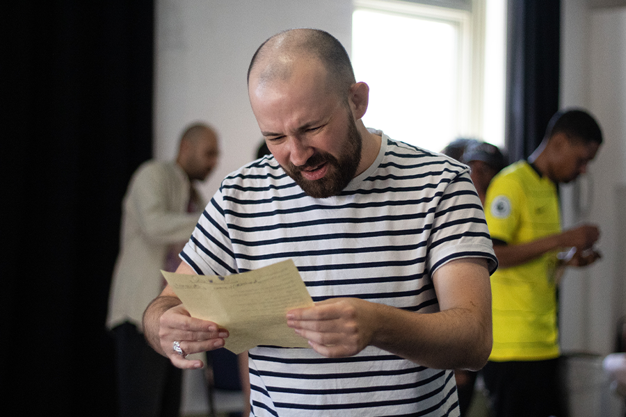 Actor Kaffe Keating holds a piece of paper looking confused, he wears a striped black and white top. He is a white man in his 30s with a beard