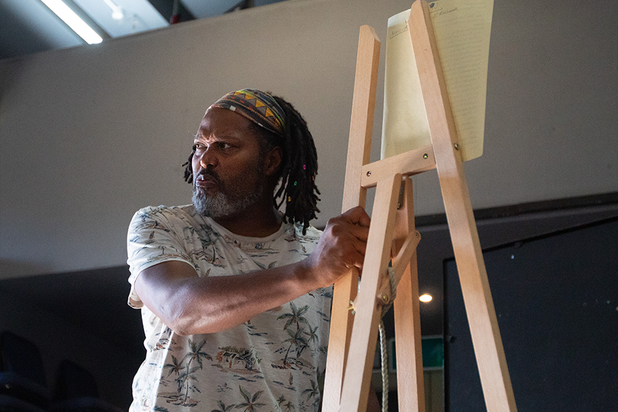Actor David Monteith holds onto an easel glaring over his shoulder. He is a black middle aged man wearing a tropical top