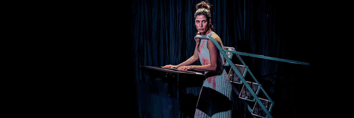 A photo of Gabriela Flarys playing the piano wearing a blue dress with red spilt down it and a blue ladder around her neck