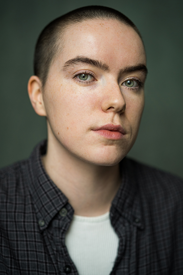 Headshot of Meg Lewis. They are caucasian and have a dark buzzcut. She wears a plaid grey shirt over a white shirt and is basing into the camera.