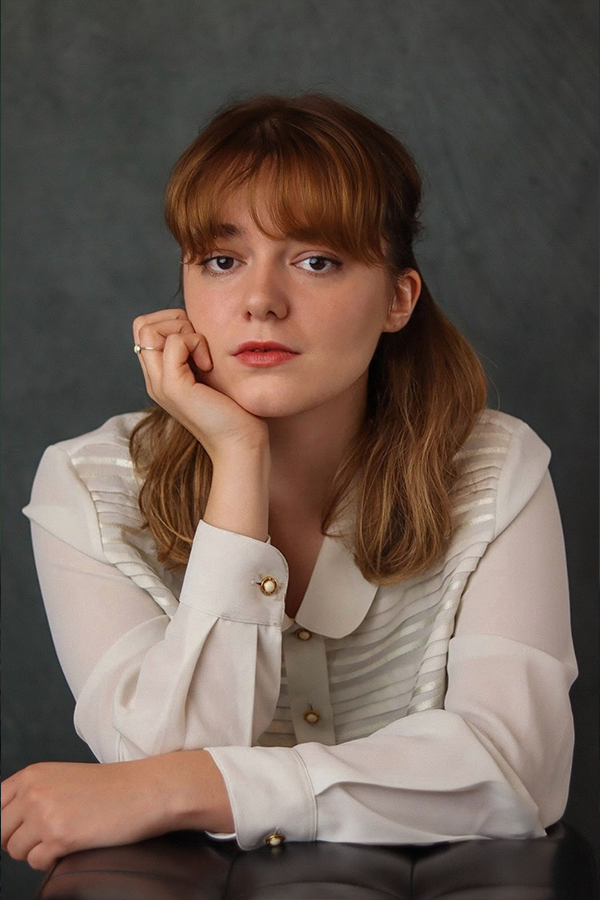 Headshot of Poppy Allen-Quarmby. She wears a white sheer shirt and holds her chin in one hand placed against her cheek.