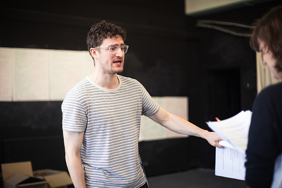 Actor Luke Nunn wears a white striped top with glasses. He is caucasian with brown curly hair. He holds a script in one hand and is talking to the other actor.