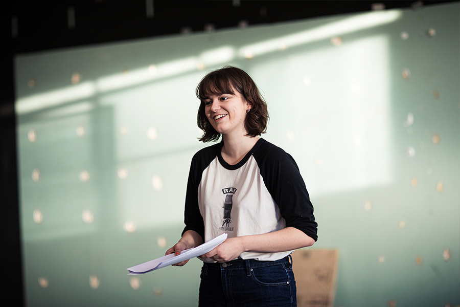 Poppy is a caucasian woman with short brown hair with a fringe. She wears a white top with black long sleeves and is holding a script smiling.