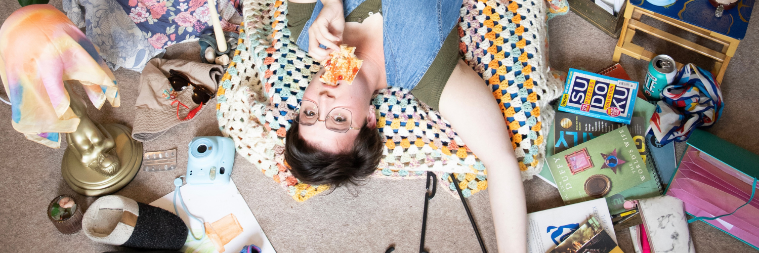 A person laying on the floor with a lot of things around them, wearing glasses and eating toast with Jam.