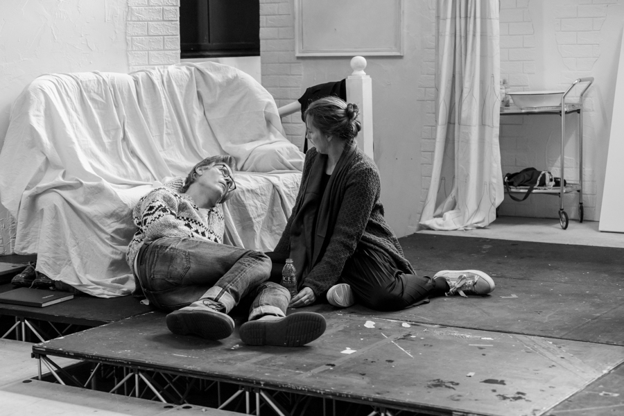 Two actors rehearsing, one lies on the floor propping their head up against a chair covered in a white sheet