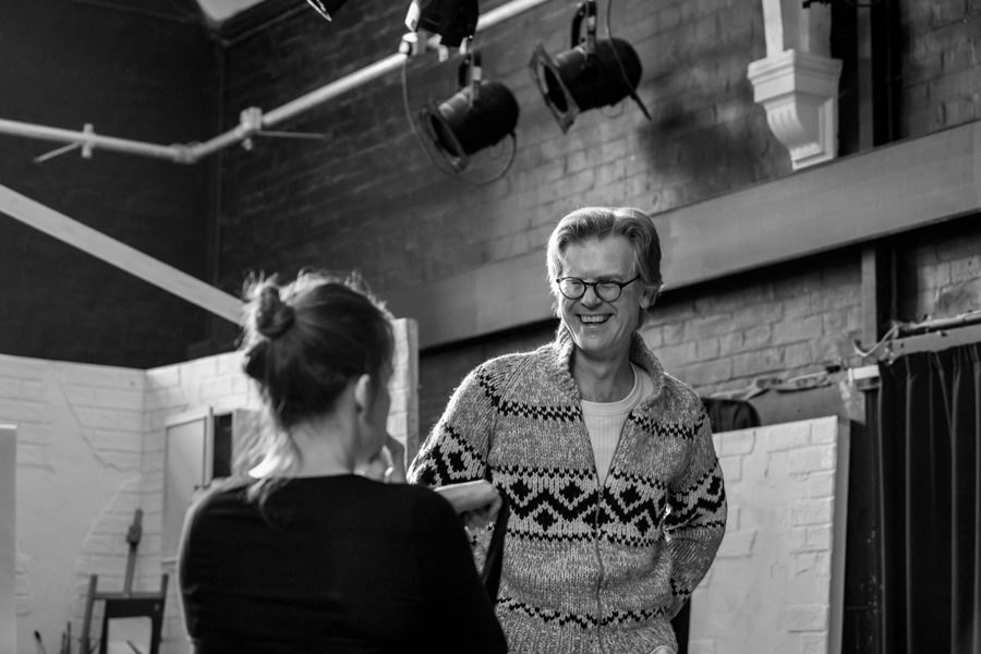 An actor wearing a patterned jumper smiles at another actor stood opposite them