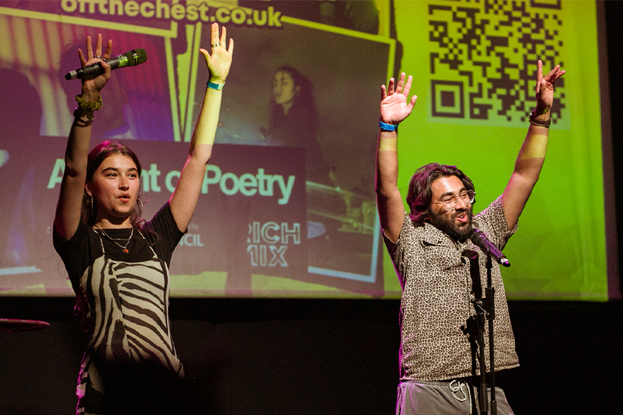 Ella and Iftikhar hosting the Off the Chest poetry night stood in front of a projector they hold their hands in the air