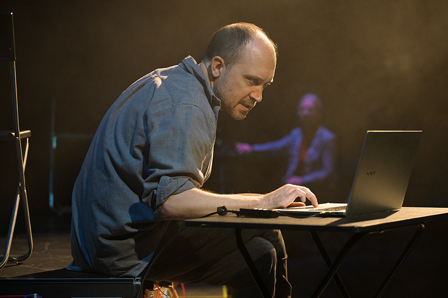 A man sits on the edge of the stage at a small table looking at a laptop screen.