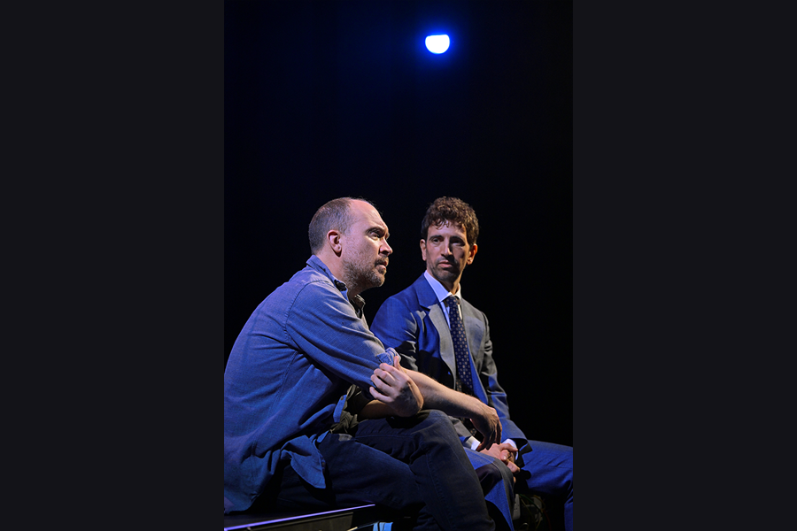 Two men sitting side by side on the edge of the stage. The man in the foreground is looking out towards the audience, the other man looks towards him.