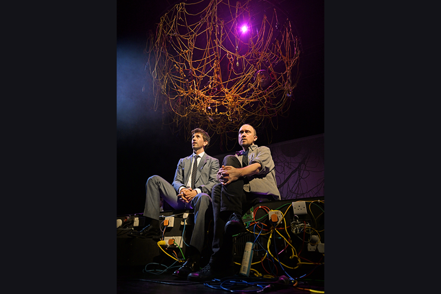 Two men sitting side by side on the edge of the stage. Both are looking out towards the audience. Around the edge of the stage is various cables and plug sockets. Above them is a structure made of cables and wires hanging from the ceiling.