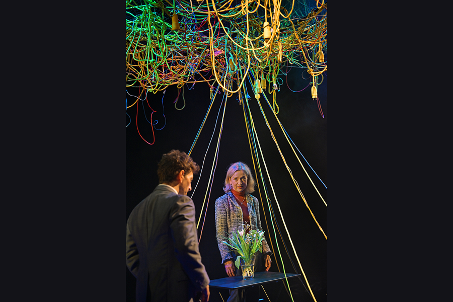A man and woman face one another across a small table which has a vase with flowers in it. The man has his back to the camera, the woman is facing the camera. Behind her and above them both is an array of cables and wires hanging down