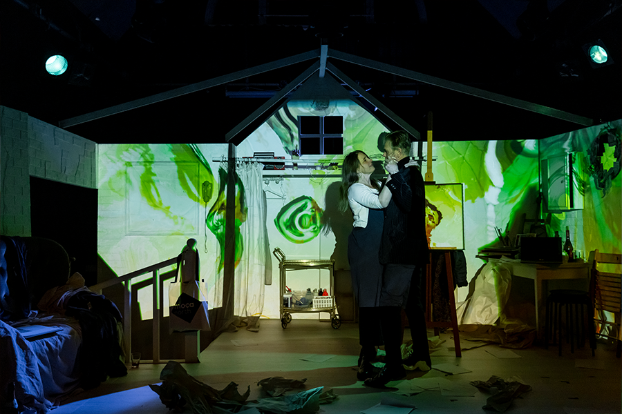 A man and woman are dance in front of a set of an art studio. The walls of the set have green patterns projected onto them.