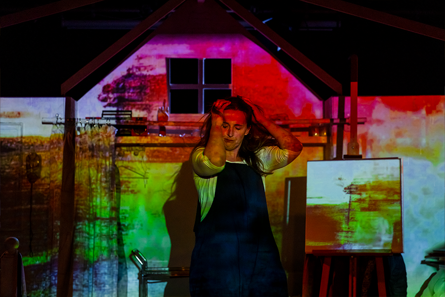 A woman moves with her hands in her hands on her head. There is a pink, green and blue colored image projected onto the wall behind her.