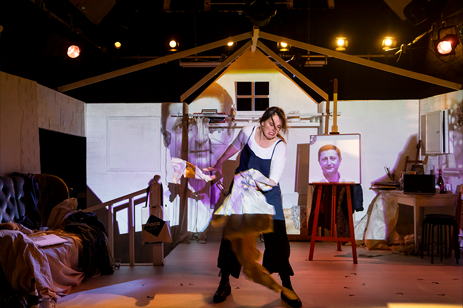 A woman aggressively rips up a large piece of paper. The background set of an art studio has projected images of a man's face.