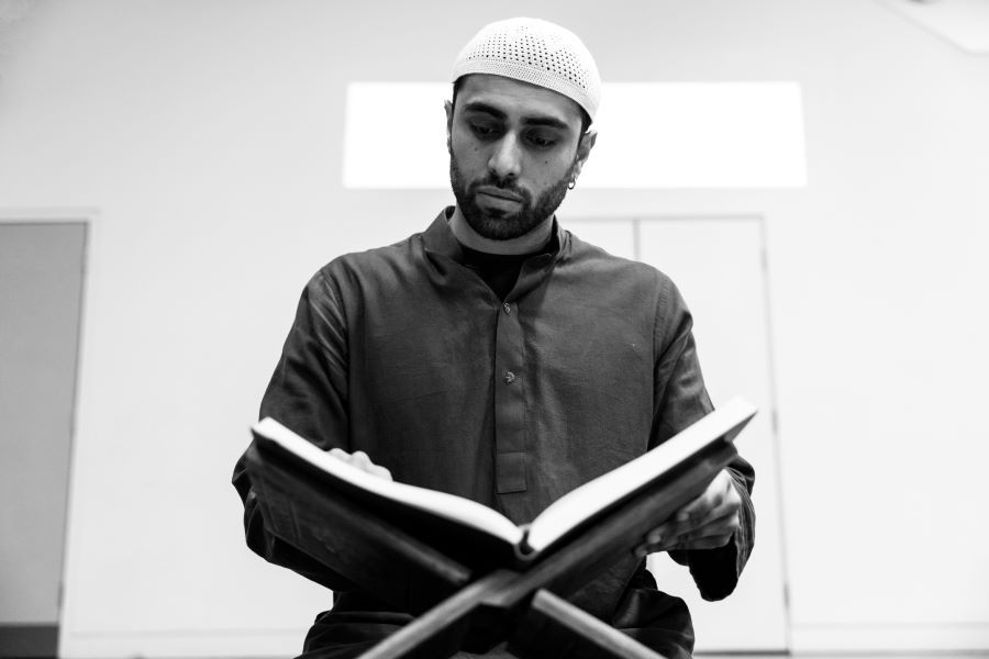 A black and white image of a man wearing a Taqiyah and reading the Quran which is propped in a book stand.