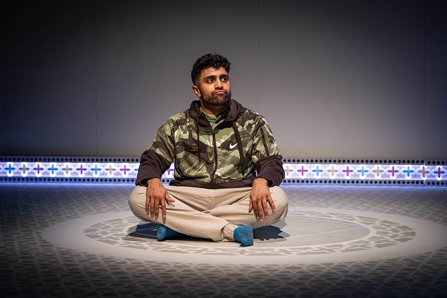 Azan Ahmed, a young south asian man, wears a camouflage nike hoodie, he is sat cross legged with blue socks.
