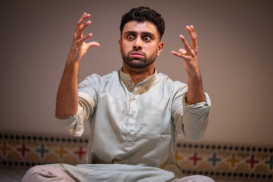 Azan Ahmed, a young south asian man, wears a Kurta, sat cross legged on the floor and holds his two hands out in front of him staring at them with wide eyes