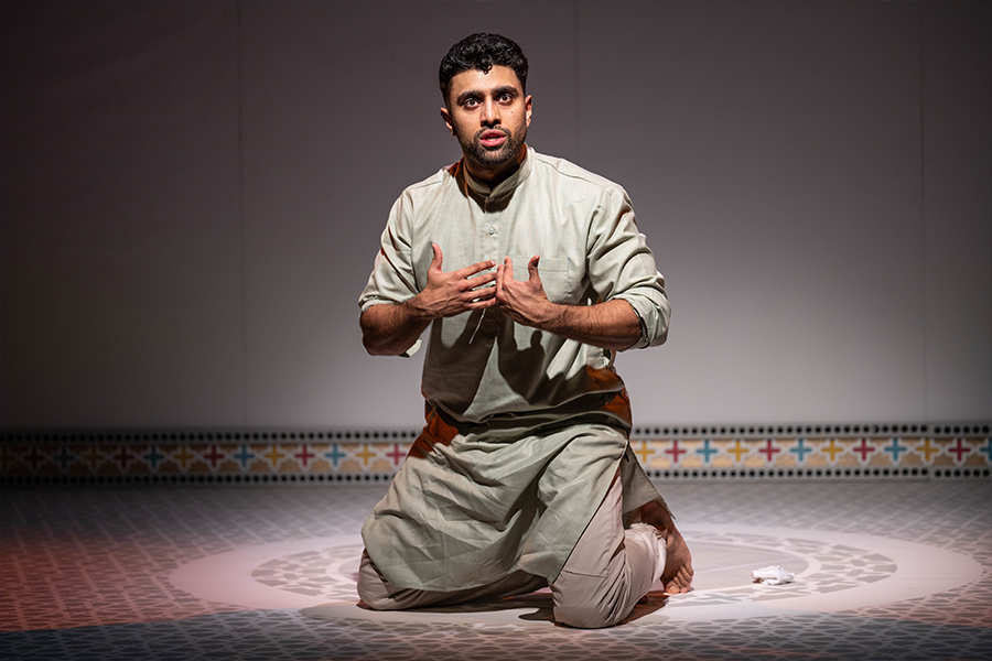 Azan Ahmed, a young south asian man, wearing a Kurta kneels on the floor with a spotlight on him. He gestures towards himself.