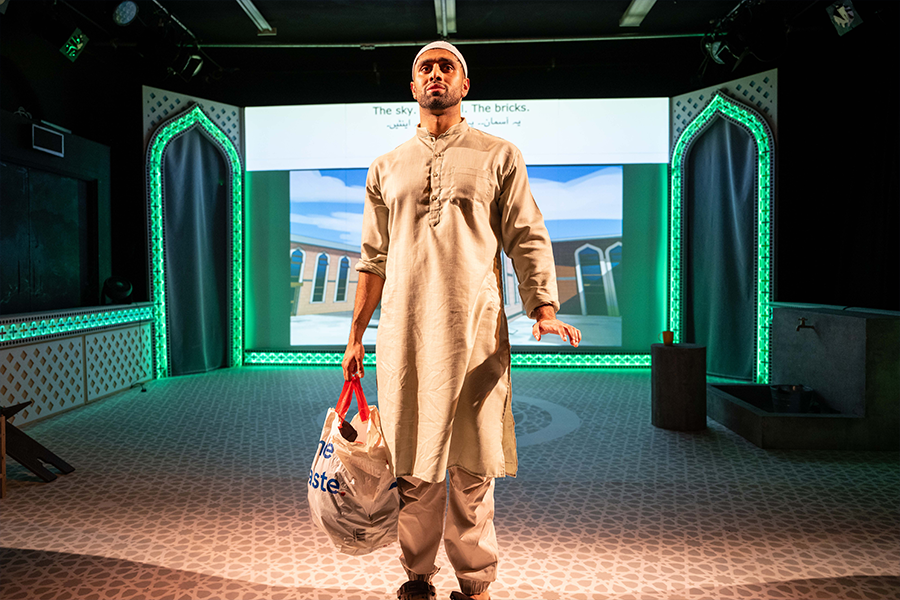 Azan Ahmed, a young south asian man, wears a Kurta and a Taqiyah. He holds a plastic Tesco bag and looks up into the audience. The stage has two door ways lit in neon green, and a mosque is projected behind him.