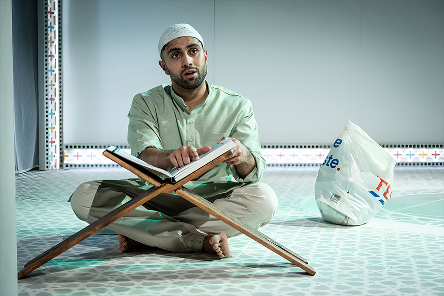 Azan Ahmed, a young south asian man, wears a Kurta and a Taqiyah. He is sat cross legged on the floor reading the Quran on a wooden Quran stand. A Tesco plastic bag sits behind him.