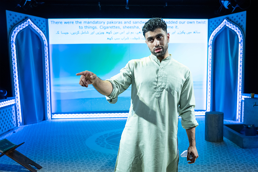 Azan Ahmed, a young south asian man, wearing a Kurta points into the crowd. There is projection behind him with captions in Urdu and English.