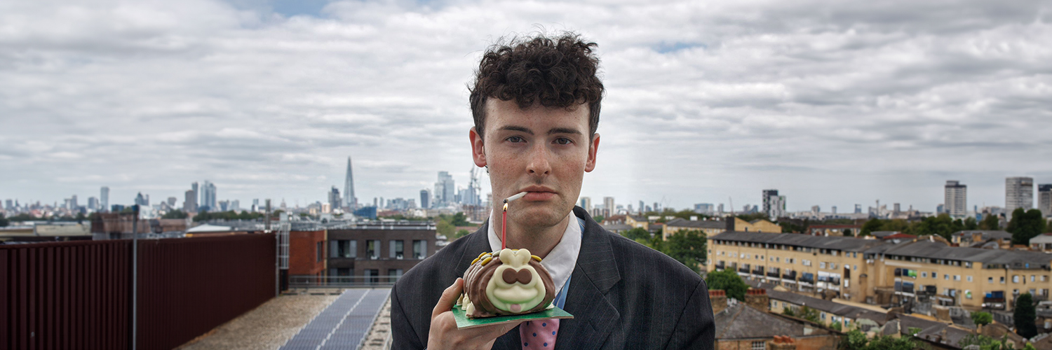 A man stands directly in the man standing center frame. We see the skyline of London behind him. He is dressed in suit. He holds up a Colin the Caterpillar cake. The candle in the cake lights up the cigarette in his mouth.