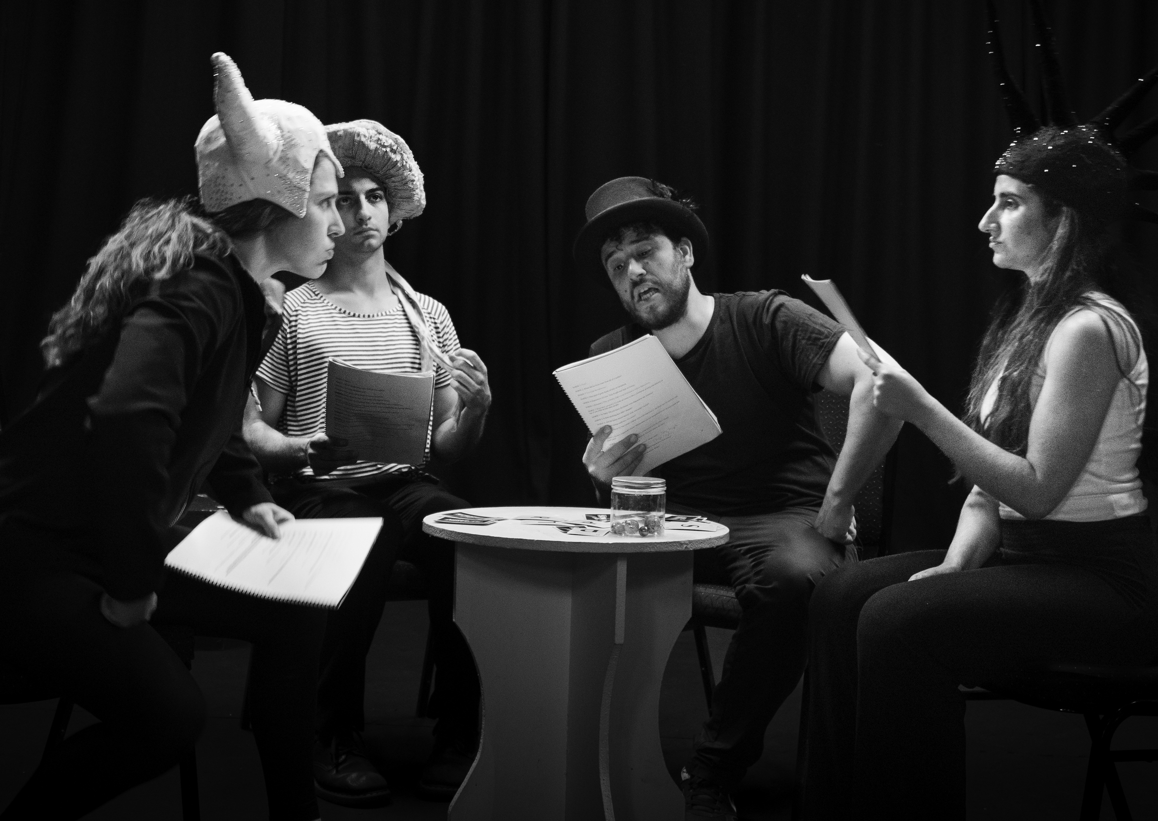 A black and white image of 4 people on stage. They all wear different hats. The woman on the far left looks across at the woman on the far right with an angry expression on her face. The man next to her on her right looks out to the middle distance. The man next to him reads from a script. And finally the last woman on the right holds a script and looks at the woman on the left.